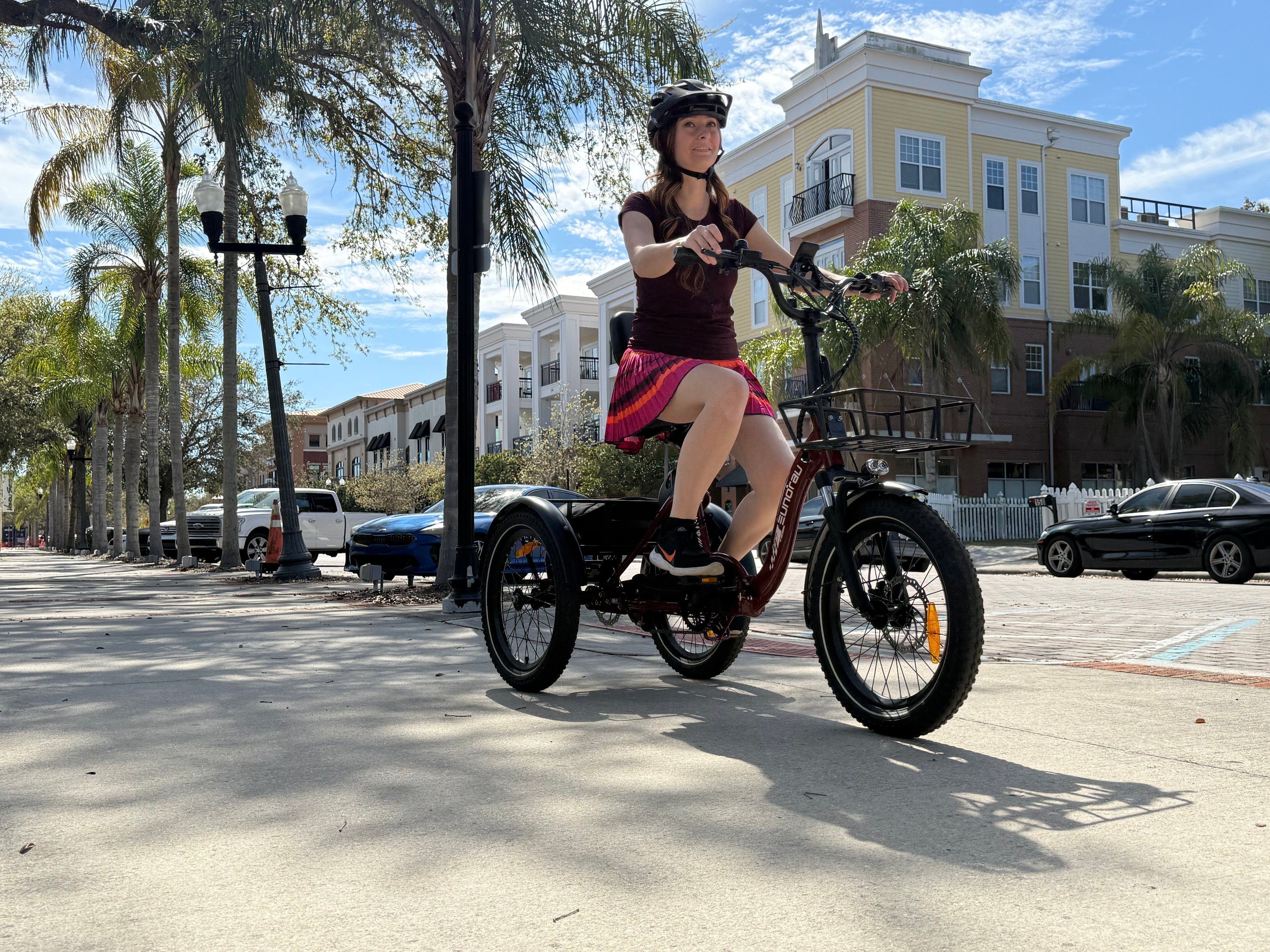 Woman riding the ONE-TRIKE 2.0 rear drive tricycle along a sunny waterfront pathway.